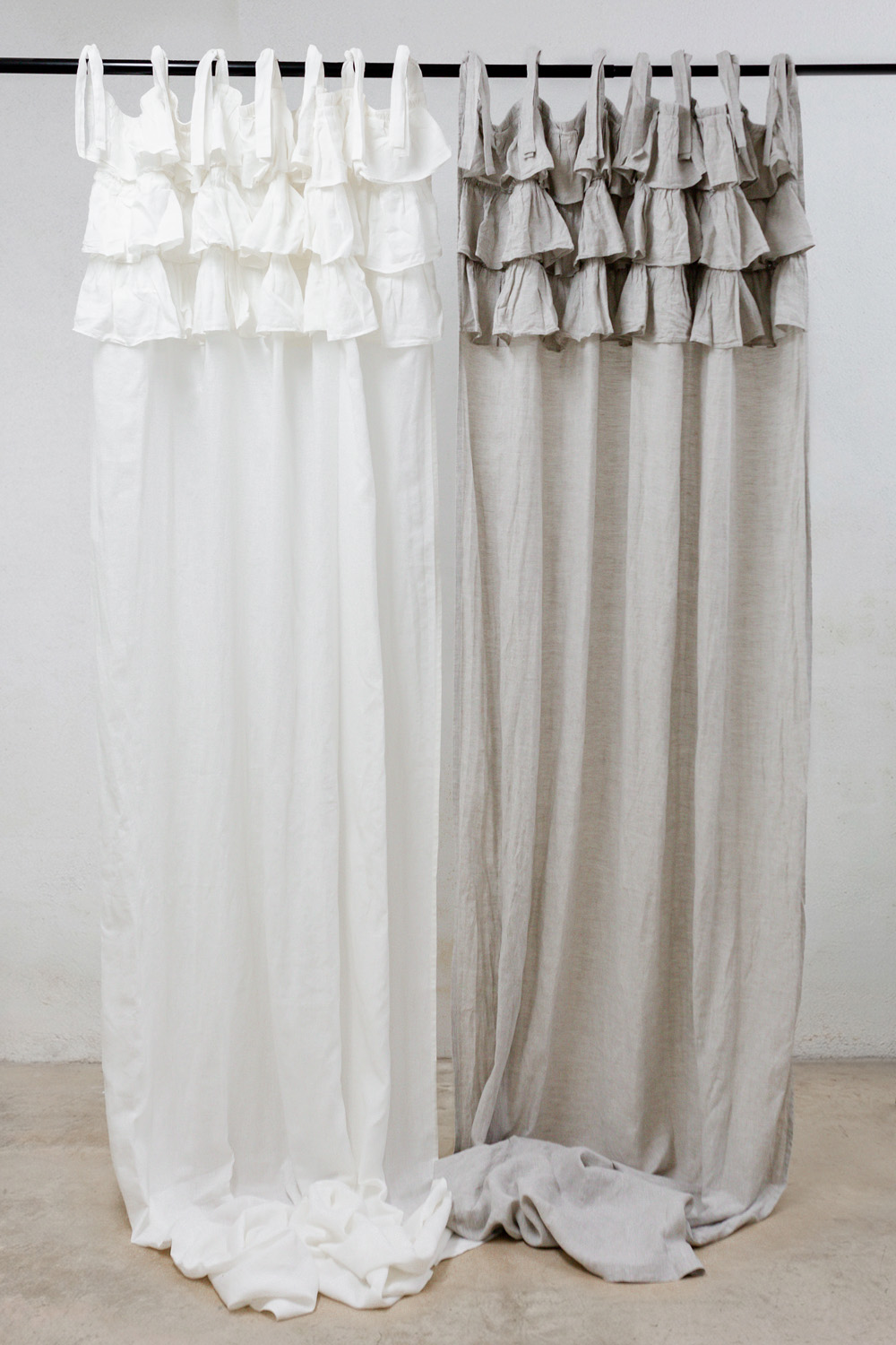 Two long curtains in white and light gray hang from a rod, with ruffled tops and fabric pooling on the floor.