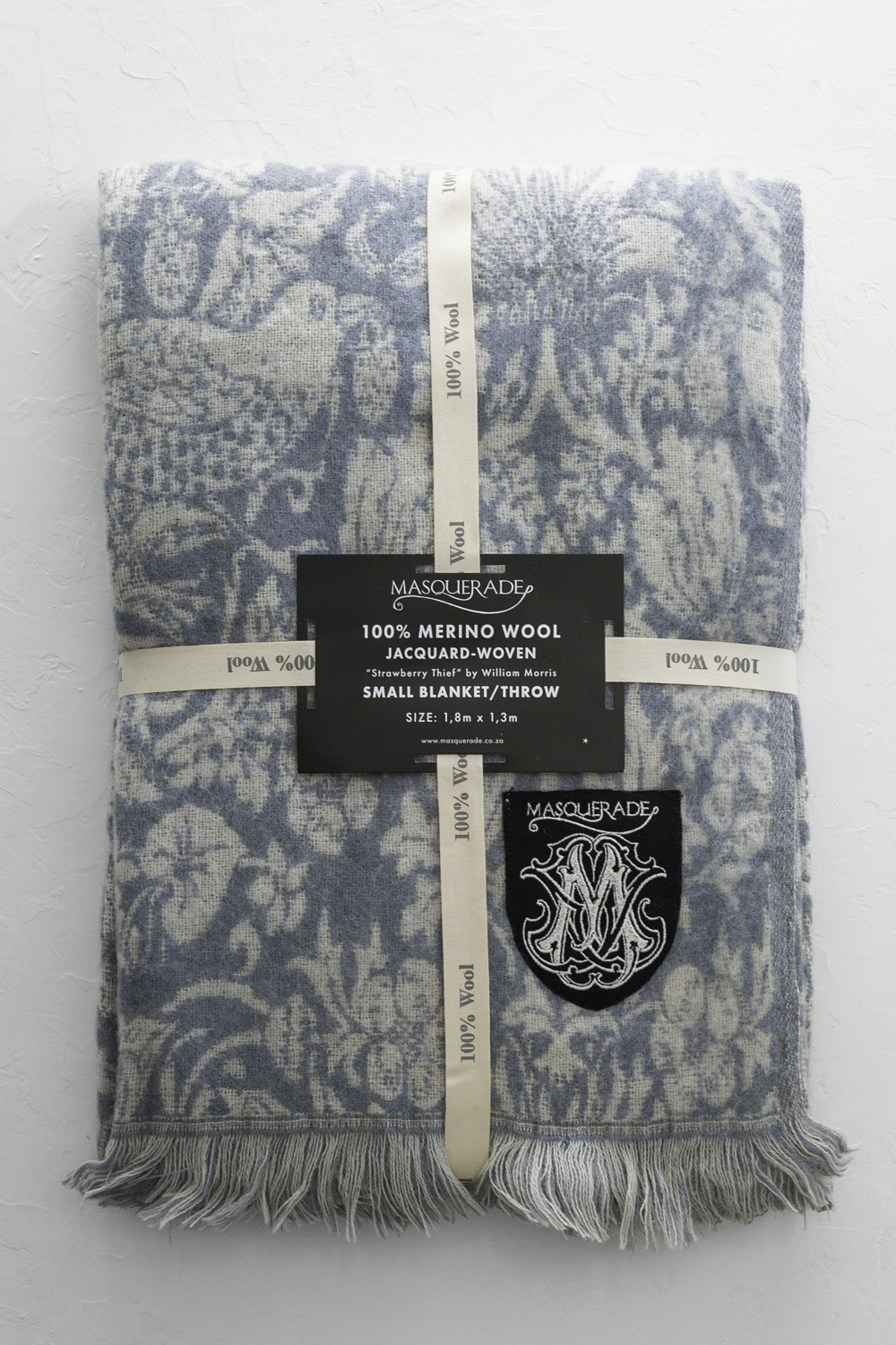 Blue jacquard-woven Merino wool throw blanket with fringe edges, wrapped with cream ribbons and a black Masquerade label and patch.