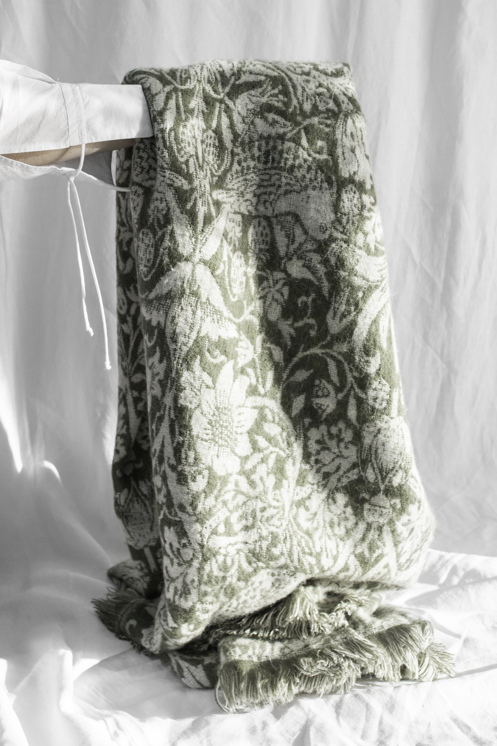 Green and white damask-pattern blanket draped over a rod, with fringed edges pooling at the bottom on a white backdrop.